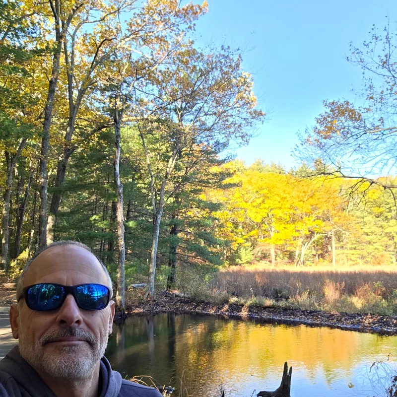 Patrick in blue sunglasses by a pond, fall trees behind him